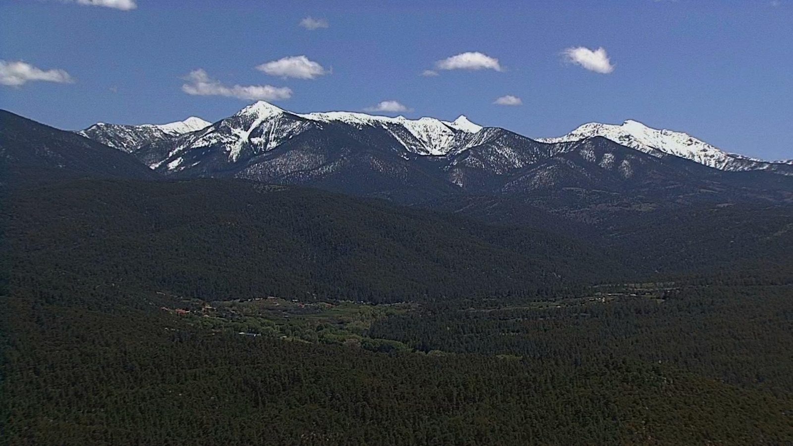 aerial photo showing green hills and El Valle, New Mexico in front of the mountains.